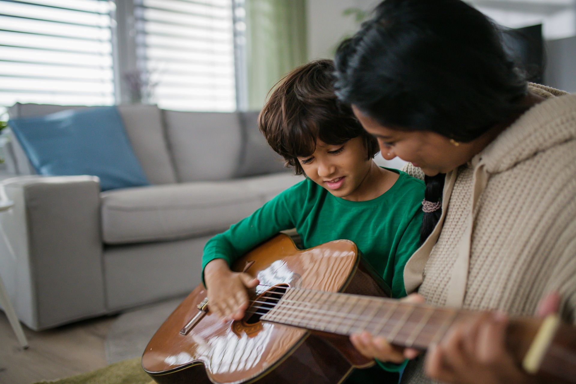 A woman and child smiling as they play an acoustic guitar together at home.